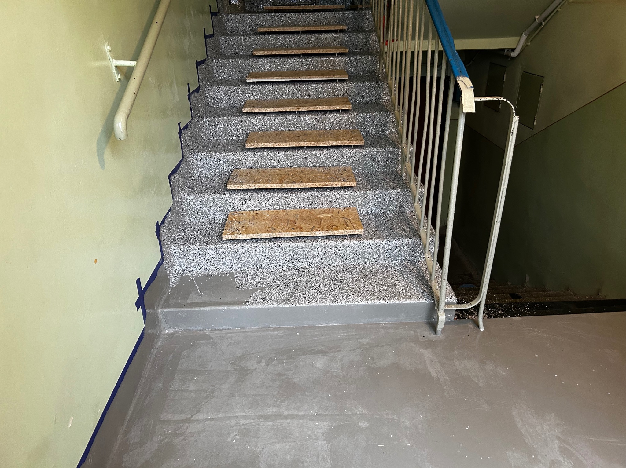 Schody w trakcie renowacji: pomalowane stopnie zabezpieczone płytami OSB, ściany z taśmą malarską. Widoczne detale wykończenia i przygotowania do dalszych prac.