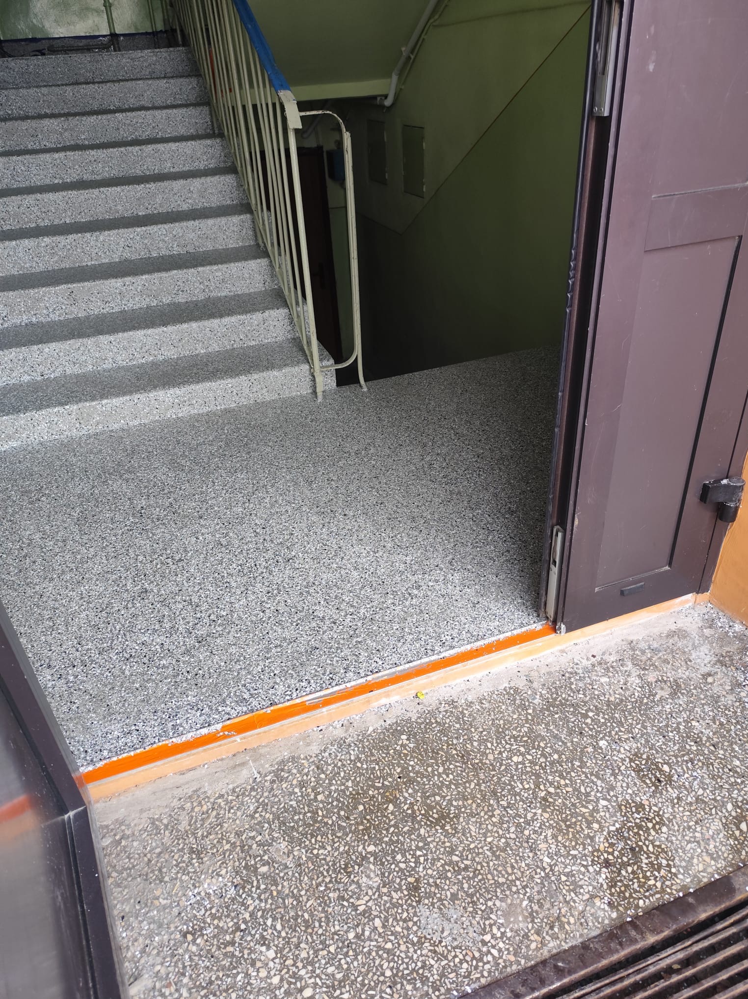 Schody i podłoga w klatce schodowej wykończone szarym lastryko. Widoczne brązowe drzwi i fragment metalowej barierki. Pomarańczowy próg.