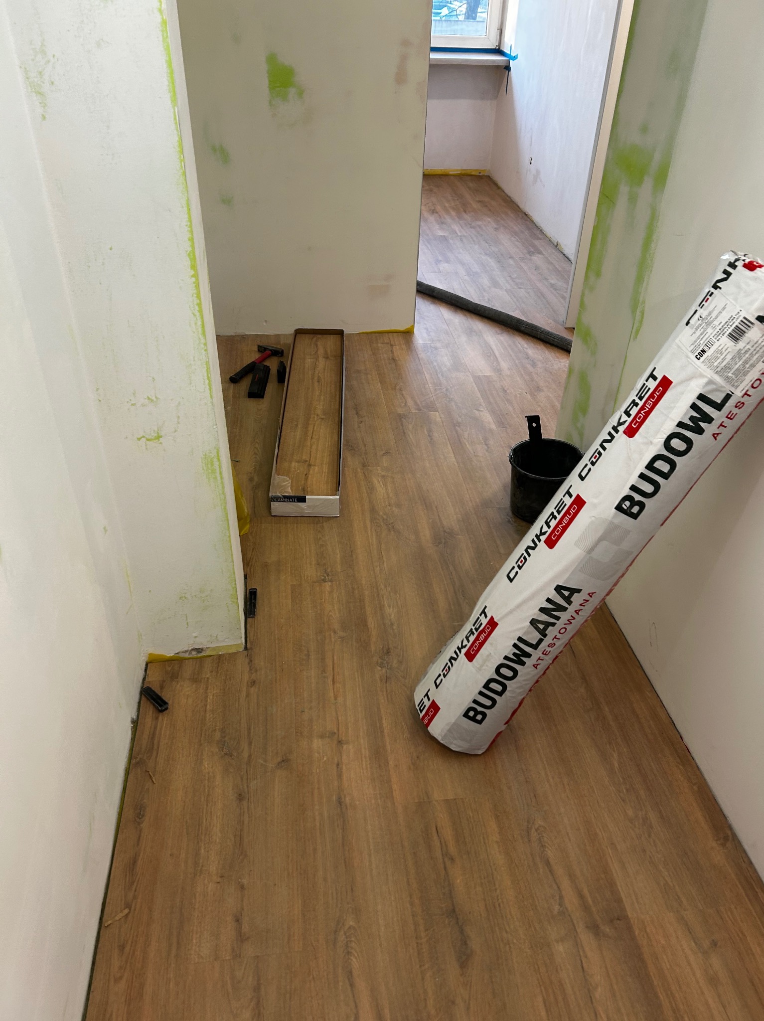 Układanie paneli podłogowych w trakcie remontu: widoczna podłoga z paneli, narzędzia, paczka paneli i rolka podkładu. Ściany przygotowane do malowania.