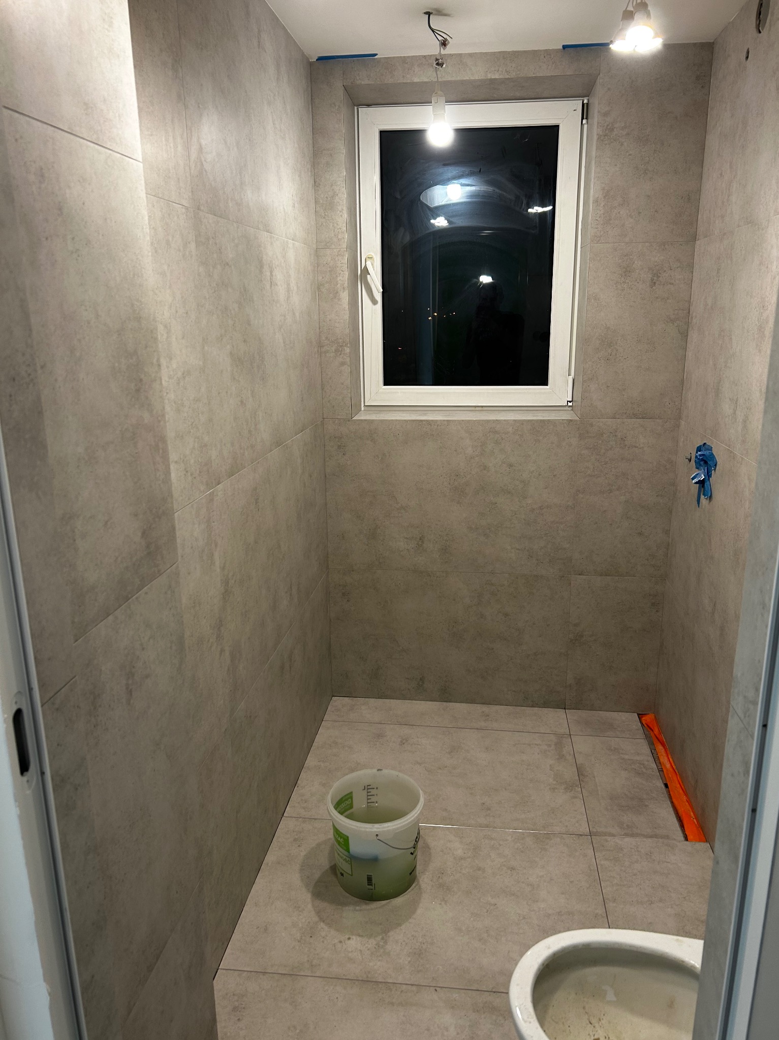 Wykończona łazienka z szarymi płytkami, widoczne okno i wiadro. Minimalistyczny design, surowe oświetlenie. Częściowo widoczna toaleta i pomarańczowa listwa.