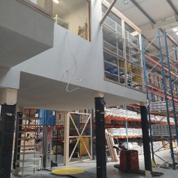 LUNAN SP. Z O.O. - Wnętrze hali magazynowej z antresolą w trakcie budowy: widoczne surowe płyty kartonowo-gipsowe, drewniane konstrukcje i metalowe regały magazynowe w tle.