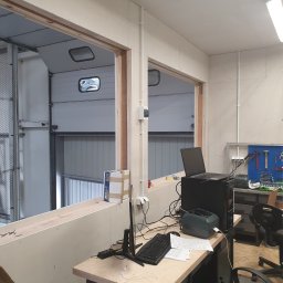 LUNAN SP. Z O.O. - Wnętrze warsztatu z widokiem na bramę garażową przez otwór w ścianie z płyt kartonowo-gipsowych, biurko z komputerem i narzędzia w tle.