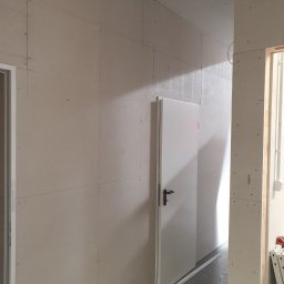 LUNAN SP. Z O.O. - Wnętrze w trakcie prac wykończeniowych: ściany obłożone płytami gipsowo-kartonowymi z widocznymi łączeniami, białe drzwi z czarną klamką i listwa przypodłogowa z otworami, widoczne ślady po ołówku...