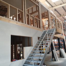 LUNAN SP. Z O.O. - Wnętrze hali w trakcie budowy z drewnianą konstrukcją szkieletową, ścianami z płyt gipsowo-kartonowych i metalowymi schodami, widoczne regały magazynowe w tle.