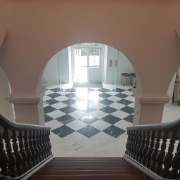 LUNAN SP. Z O.O. - Widok z góry schodów na hol z czarno-białą marmurową posadzką w szachownicę, z białymi ścianami, łukowym przejściem i przeszklonymi drzwiami wejściowymi w tle.