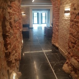 LUNAN SP. Z O.O. - Wnętrze z odrestaurowanymi ceglanymi łukami, czarną marmurową podłogą z białymi żyłami i nowoczesnym oświetleniem LED, widok na jasne pomieszczenie z przeszklonymi drzwiami.
