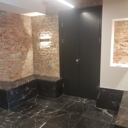LUNAN SP. Z O.O. - Wnętrze z odrestaurowaną ceglaną ścianą, czarnymi drzwiami i marmurową posadzką z białymi żyłami, oświetlenie punktowe podkreśla fakturę cegły.