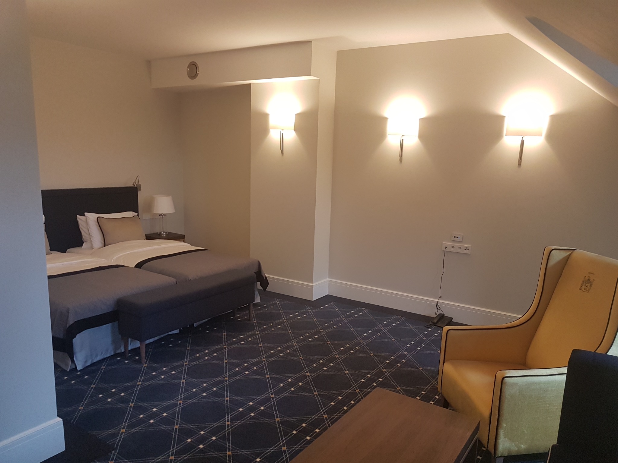 Pokój hotelowy na poddaszu z dwoma łóżkami pojedynczymi, pufą, żółtym fotelem z herbem, trzema kinkietami ściennymi i wzorzystą wykładziną dywanową.