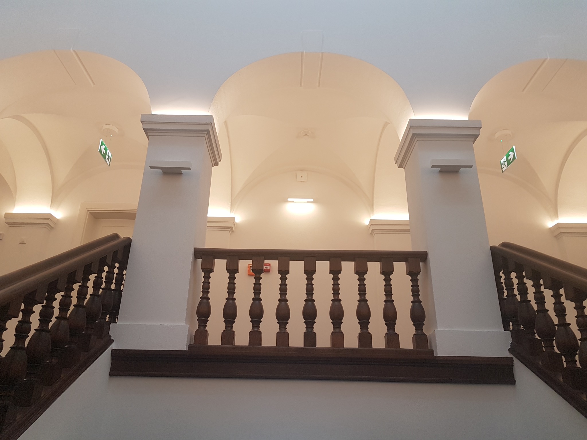 Klatka schodowa z drewnianą balustradą i białymi ścianami, widziana z dołu, z widocznymi sklepieniami łukowymi i oświetleniem LED.