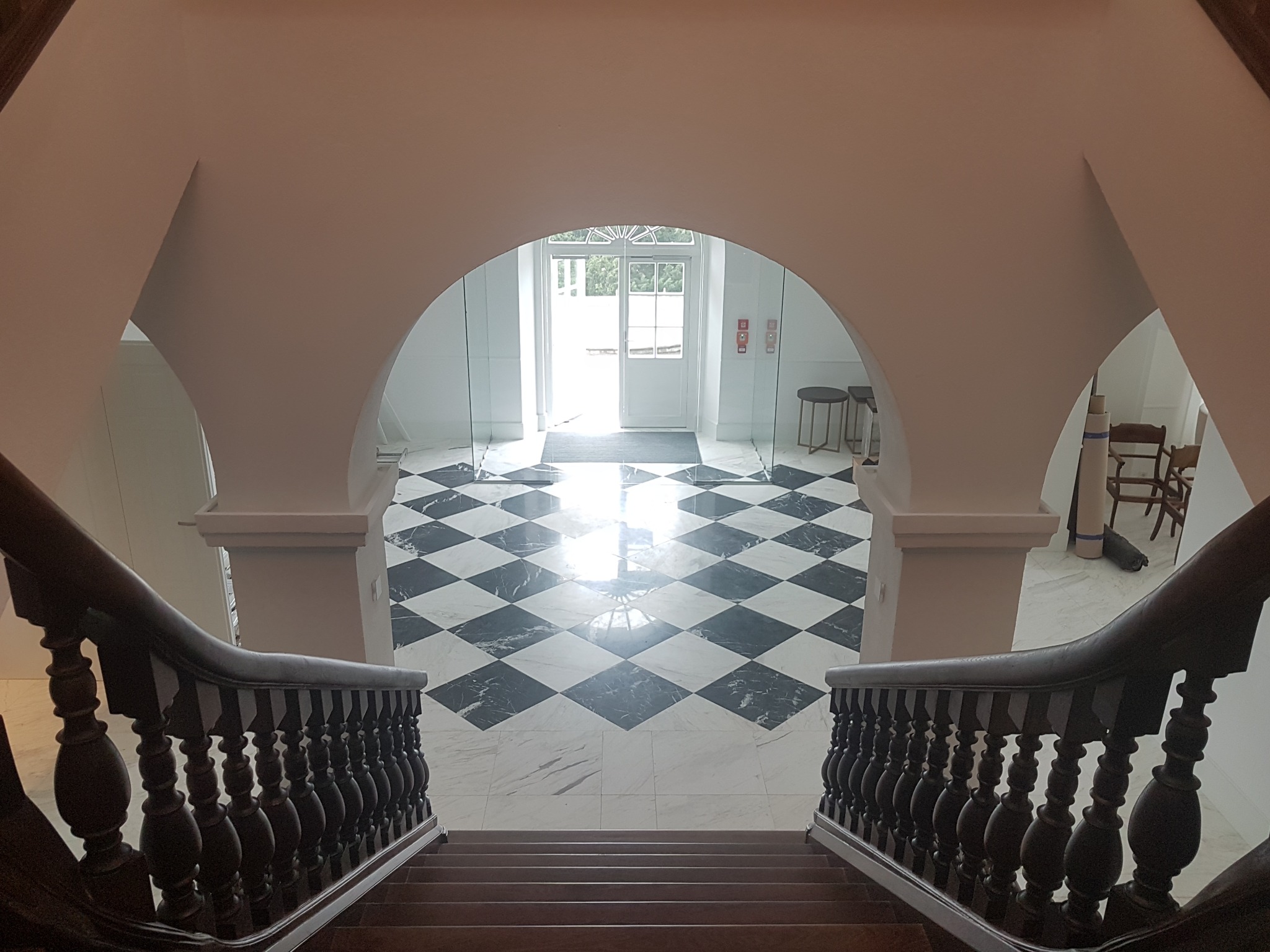 Widok z góry schodów na hol z czarno-białą marmurową posadzką w szachownicę, z białymi ścianami, łukowym przejściem i przeszklonymi drzwiami wejściowymi w tle.