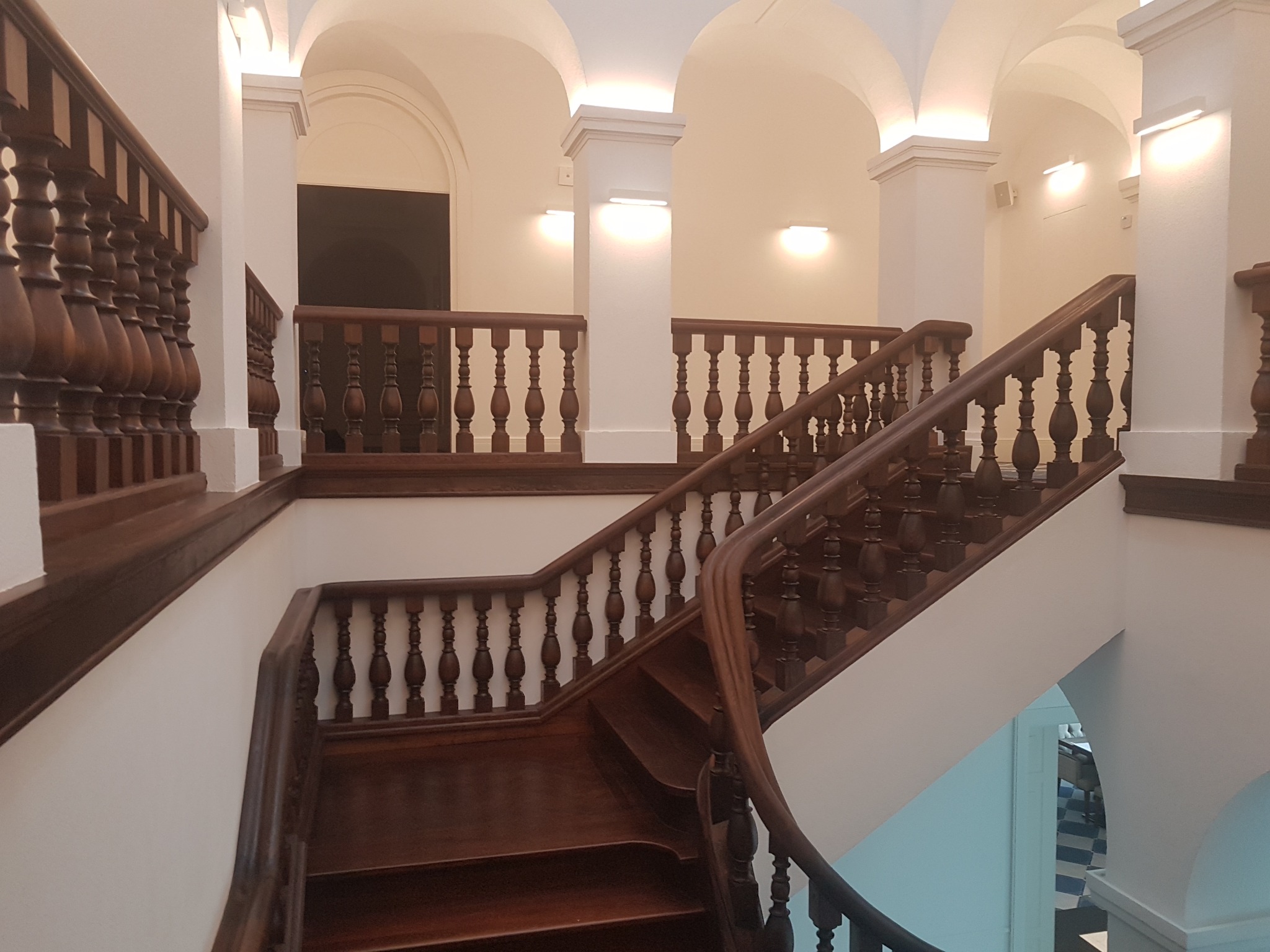 Klatka schodowa w kamienicy z drewnianymi schodami i balustradą, białe ściany i kolumny, łukowe przejścia, widoczne oświetlenie punktowe.