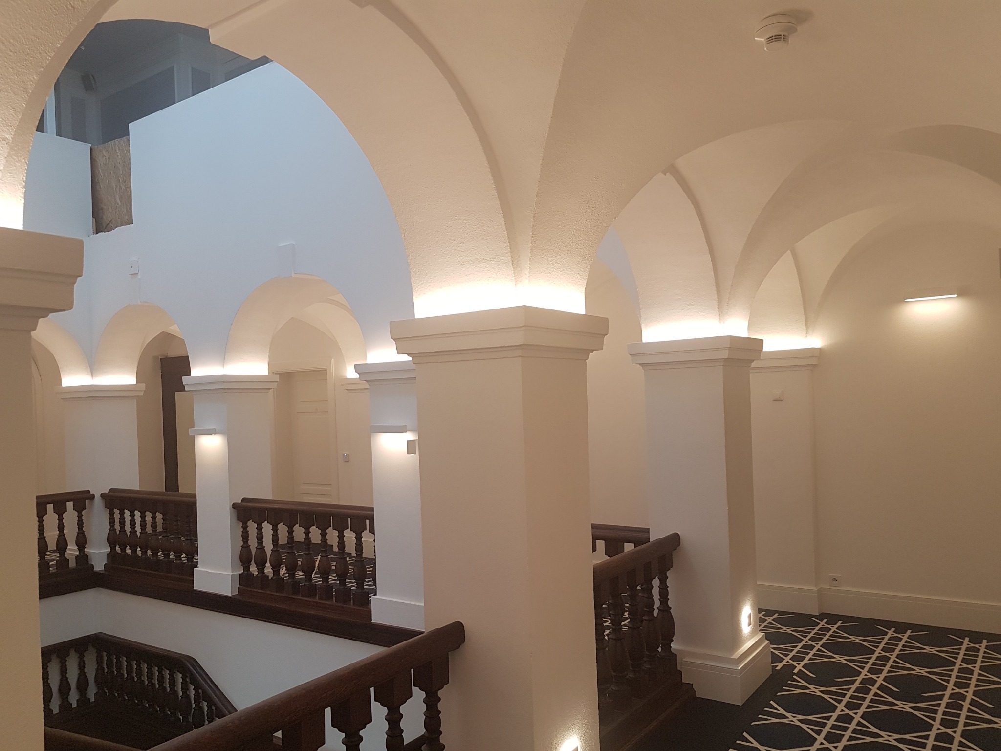 Korytarz w kamienicy z białymi łukami, kolumnami i drewnianymi balustradami, oświetlony punktowo, z wzorzystą wykładziną na podłodze.