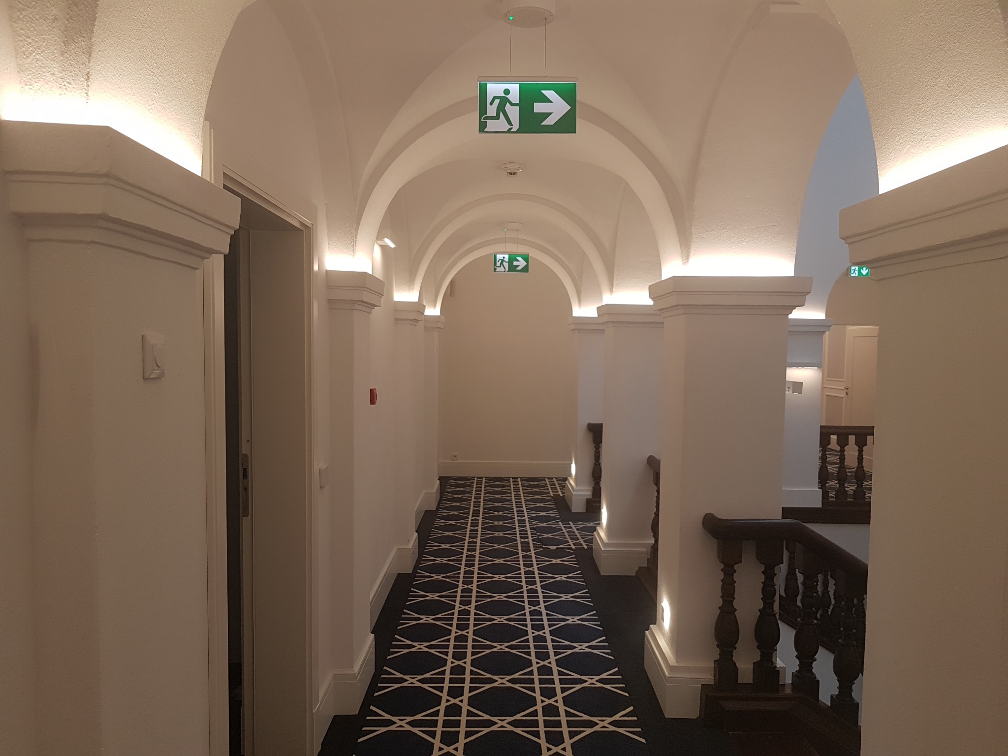 Korytarz w kamienicy z łukowym sufitem, oświetleniem LED wzdłuż ścian i wzorzystą wykładziną podłogową, zakończony schodami z drewnianą balustradą, z widocznymi znakami ewakuacyjnymi.