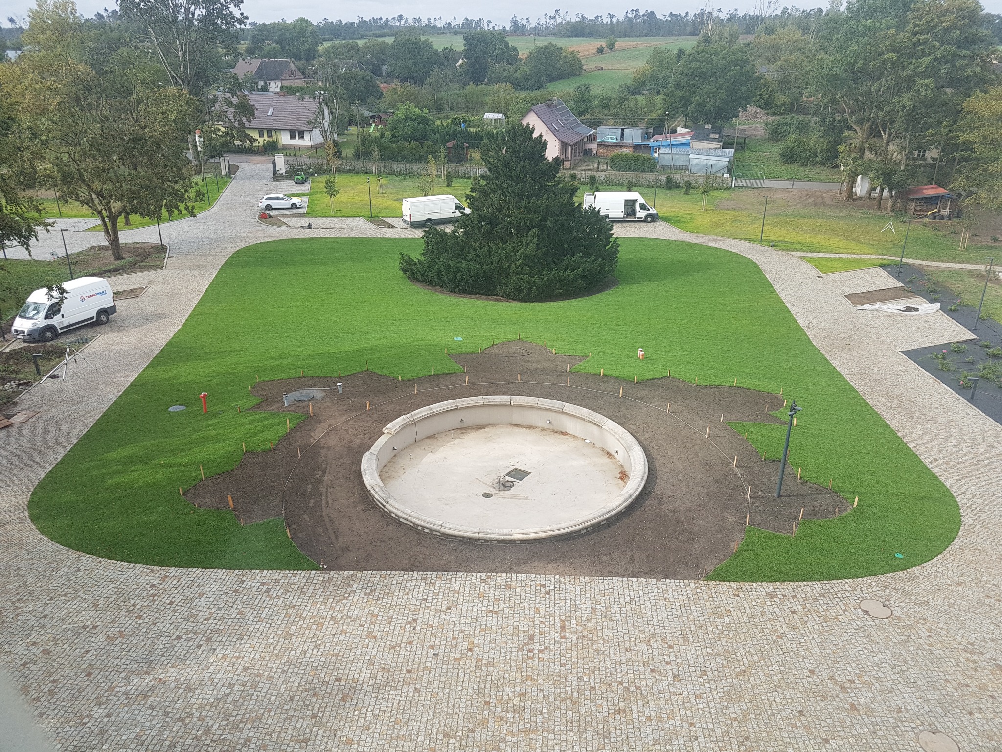 Widok z góry na plac z fontanną w trakcie budowy, świeżo położona trawa, w tle drzewa i budynki, zaparkowane białe samochody dostawcze.