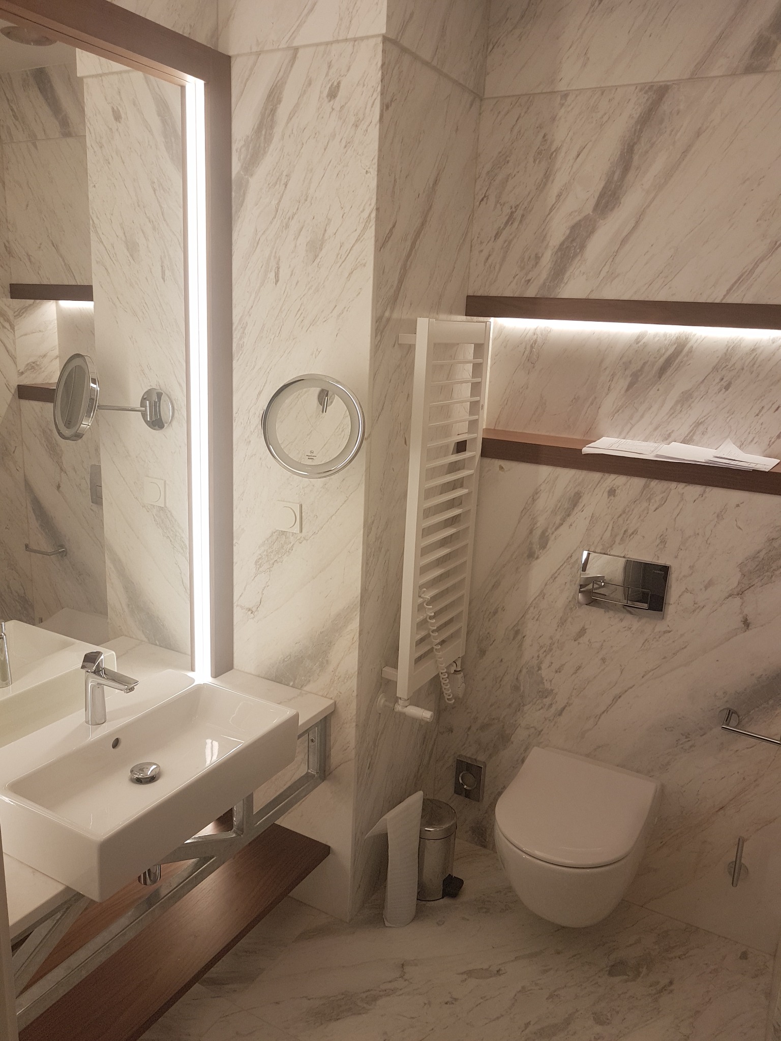 Nowoczesna łazienka wykończona marmurem, z podświetlanym lustrem, wiszącą toaletą i grzejnikiem drabinkowym.
