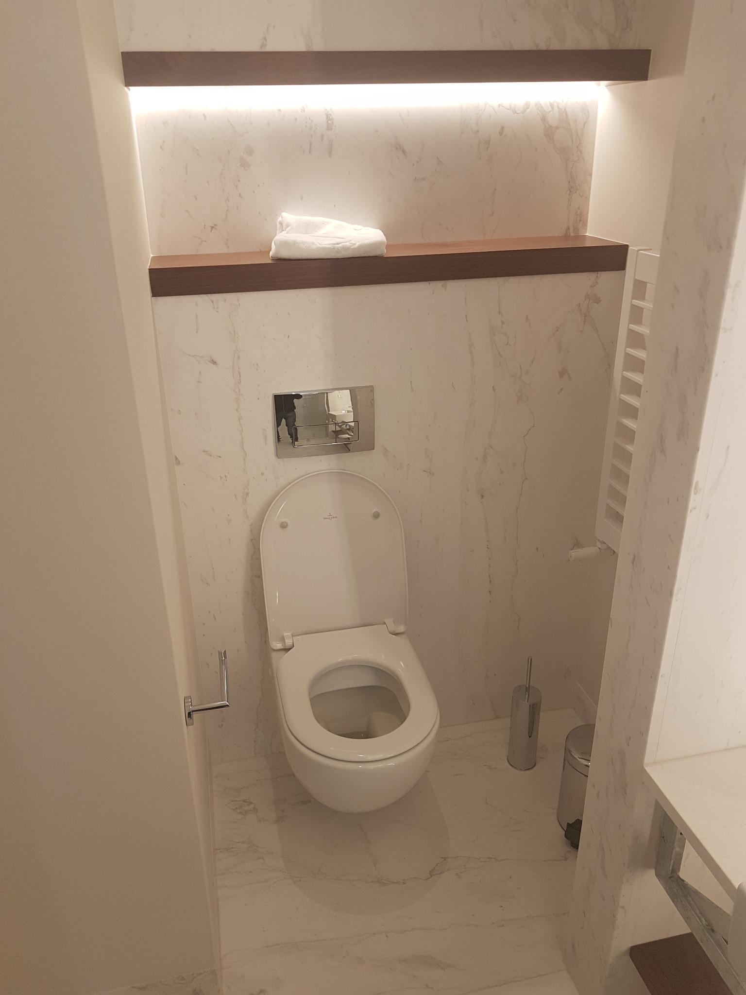 Nowoczesna toaleta Villeroy & Boch w łazience wykończonej marmurem, z drewnianymi półkami podświetlonymi LED i grzejnikiem drabinkowym.