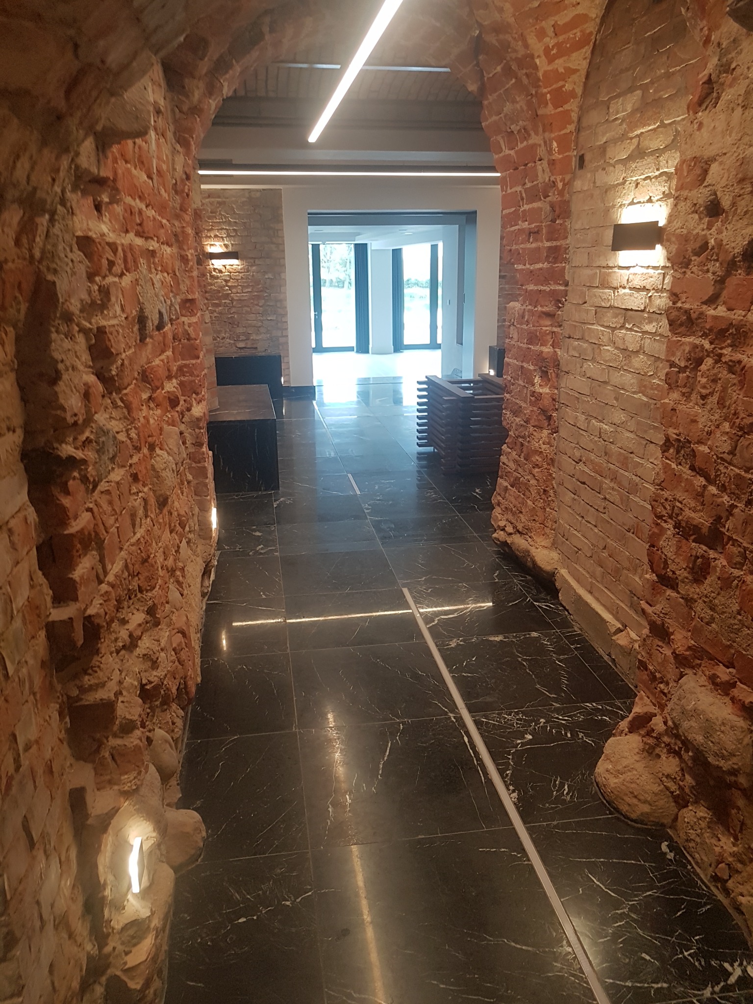 Wnętrze z odrestaurowanymi ceglanymi łukami, czarną marmurową podłogą z białymi żyłami i nowoczesnym oświetleniem LED, widok na jasne pomieszczenie z przeszklonymi drzwiami.