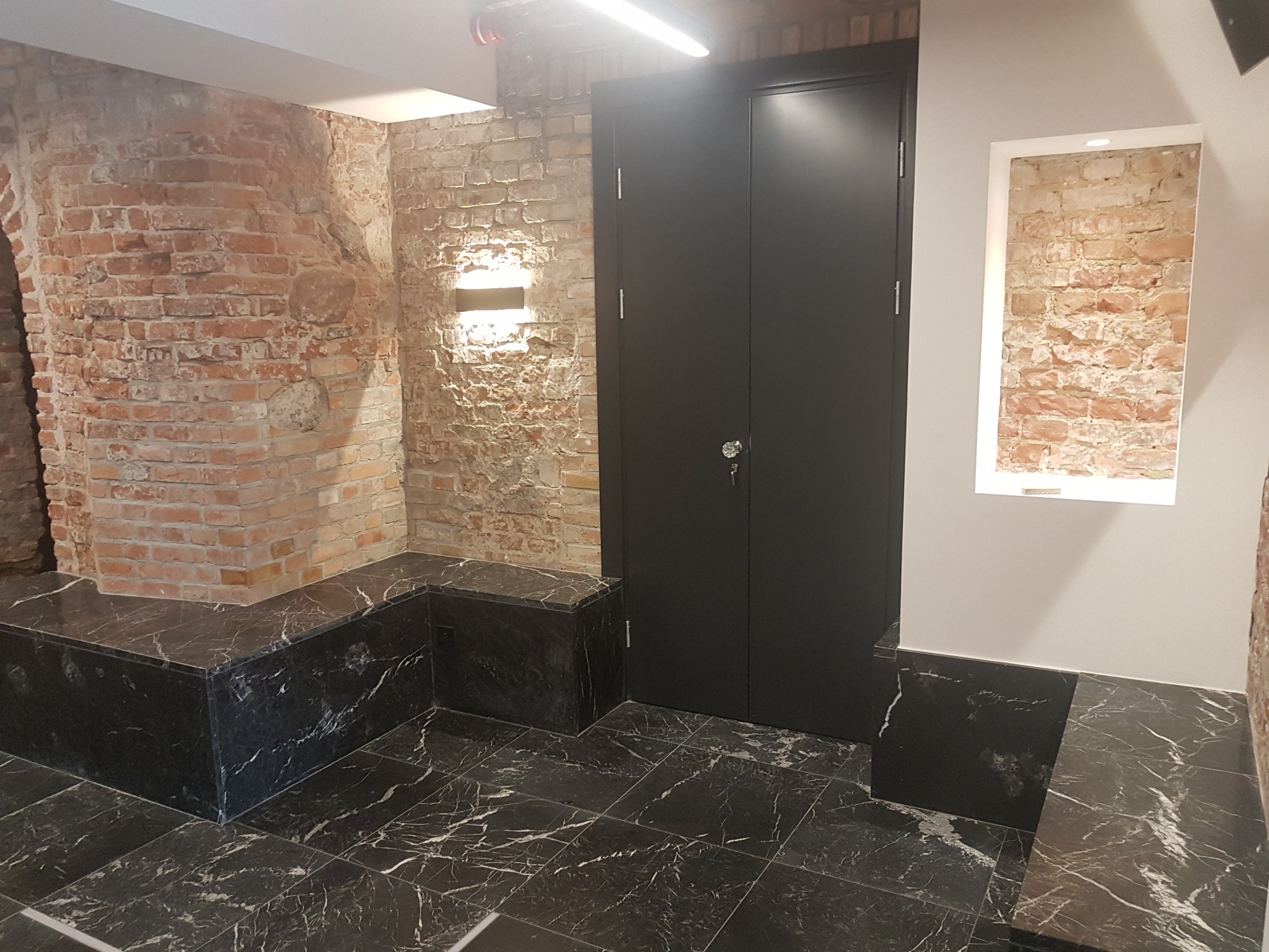 Wnętrze z odrestaurowaną ceglaną ścianą, czarnymi drzwiami i marmurową posadzką z białymi żyłami, oświetlenie punktowe podkreśla fakturę cegły.