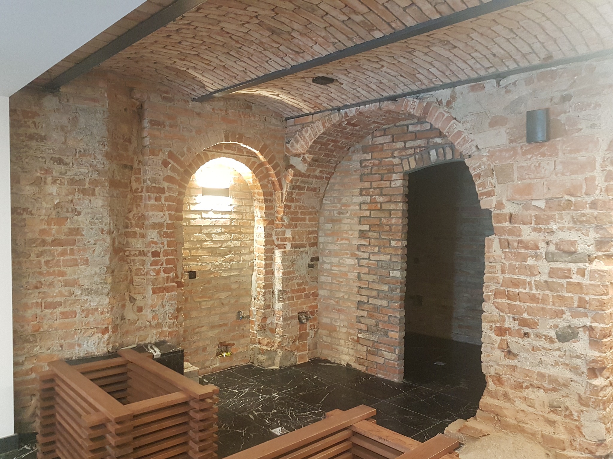 Piwnica z ceglanym sklepieniem łukowym, czarną marmurową podłogą i stosem drewnianych ram okiennych, w trakcie prac wykończeniowych.