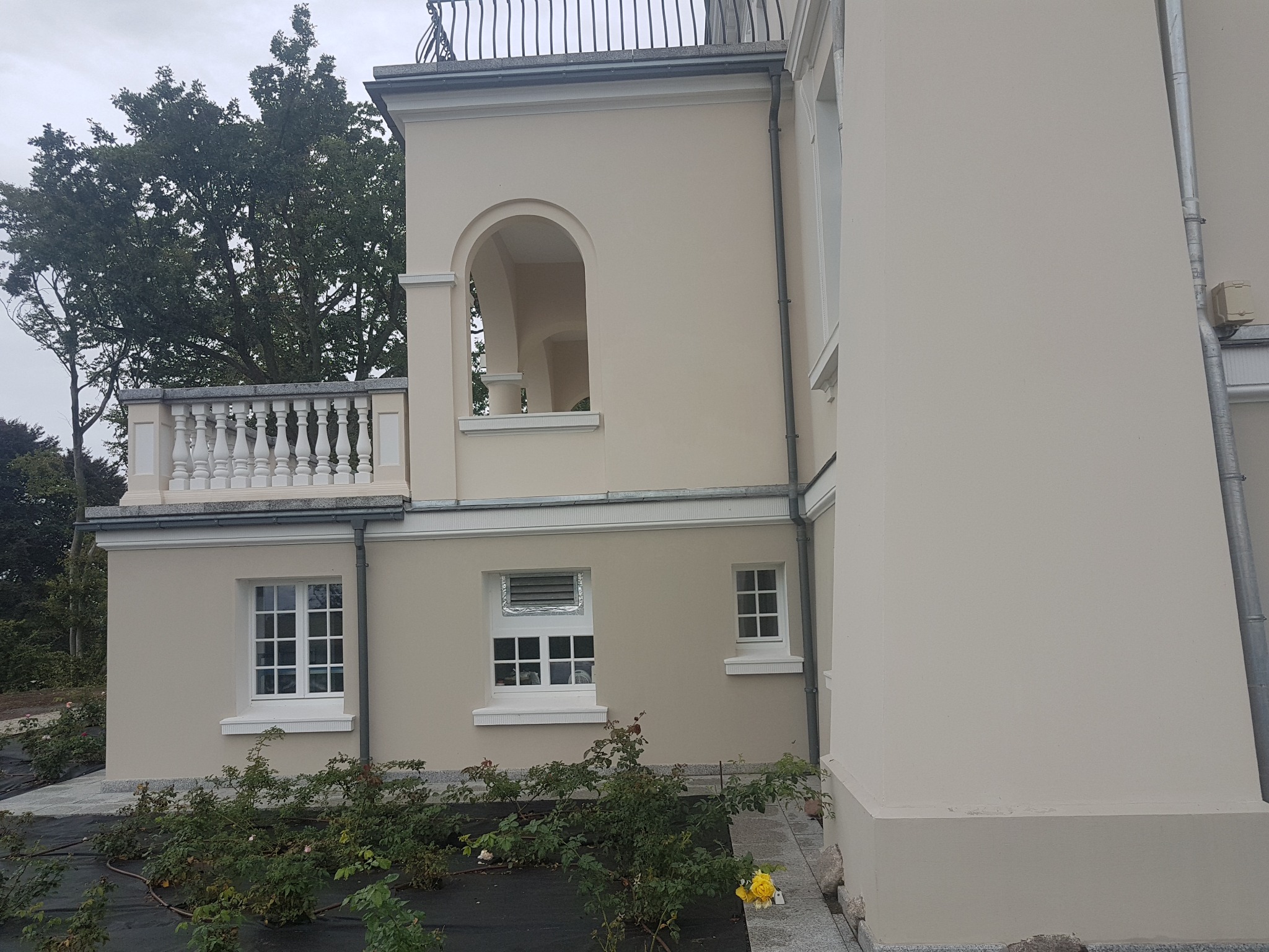 Elewacja odnowionego budynku z beżową fasadą, widoczne okna z białymi ramami, balkon z balustradą i fragment ogrodu z krzewami róż, rynna odprowadzająca wodę.