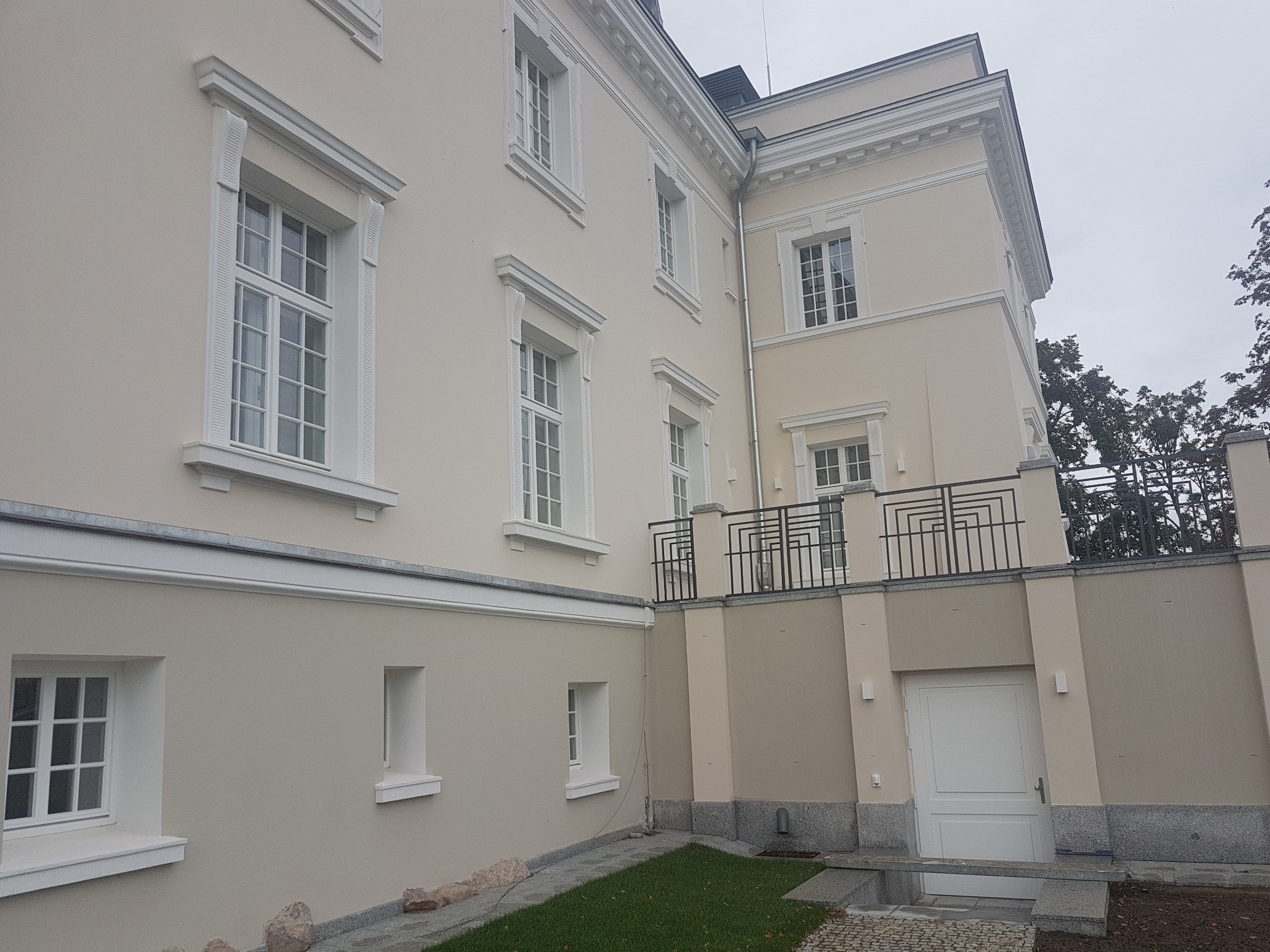 Odnowiona elewacja dwupiętrowego budynku z jasną fasadą, białymi ramami okiennymi i ozdobnymi gzymsami, widok z poziomu gruntu na fragment ściany z oknami, małym trawnikiem i fragmentem chodnika.