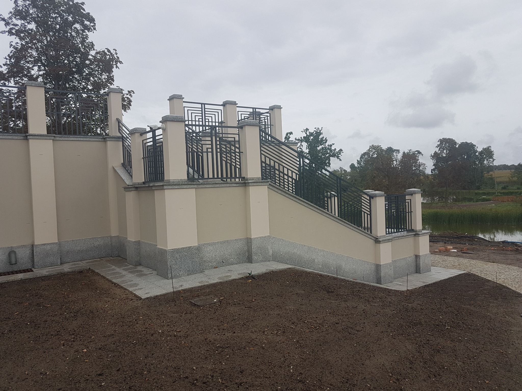 Zewnętrzne schody z balustradą w stylu art deco, prowadzące na wyższą kondygnację budynku, z widocznym fragmentem krajobrazu z jeziorem w tle, w pochmurny dzień.