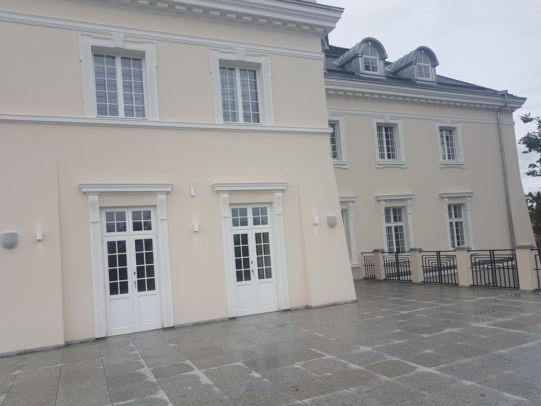 Elewacja dwukondygnacyjnego budynku z kremową fasadą, białymi oknami i drzwiami oraz dekoracyjnymi sztukateriami, widoczna z poziomu wyłożonego szarą kostką brukową.