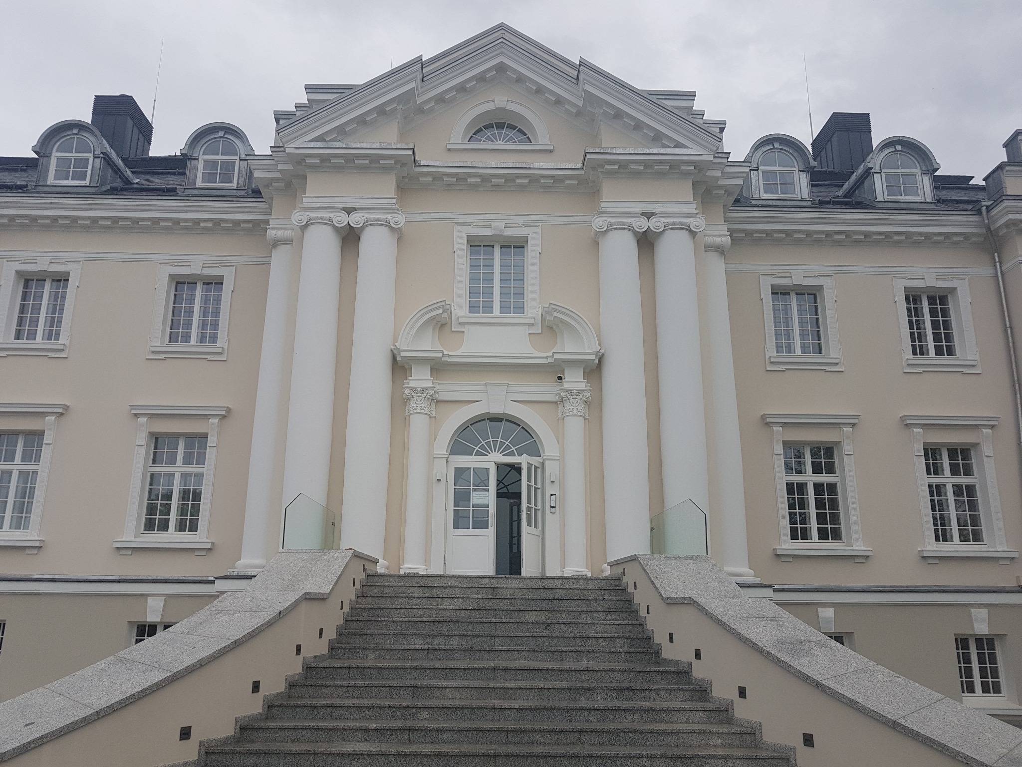 Fasada odnowionego, klasycystycznego budynku z kolumnami i otwartymi, białymi drzwiami wejściowymi, widok od dołu schodów.