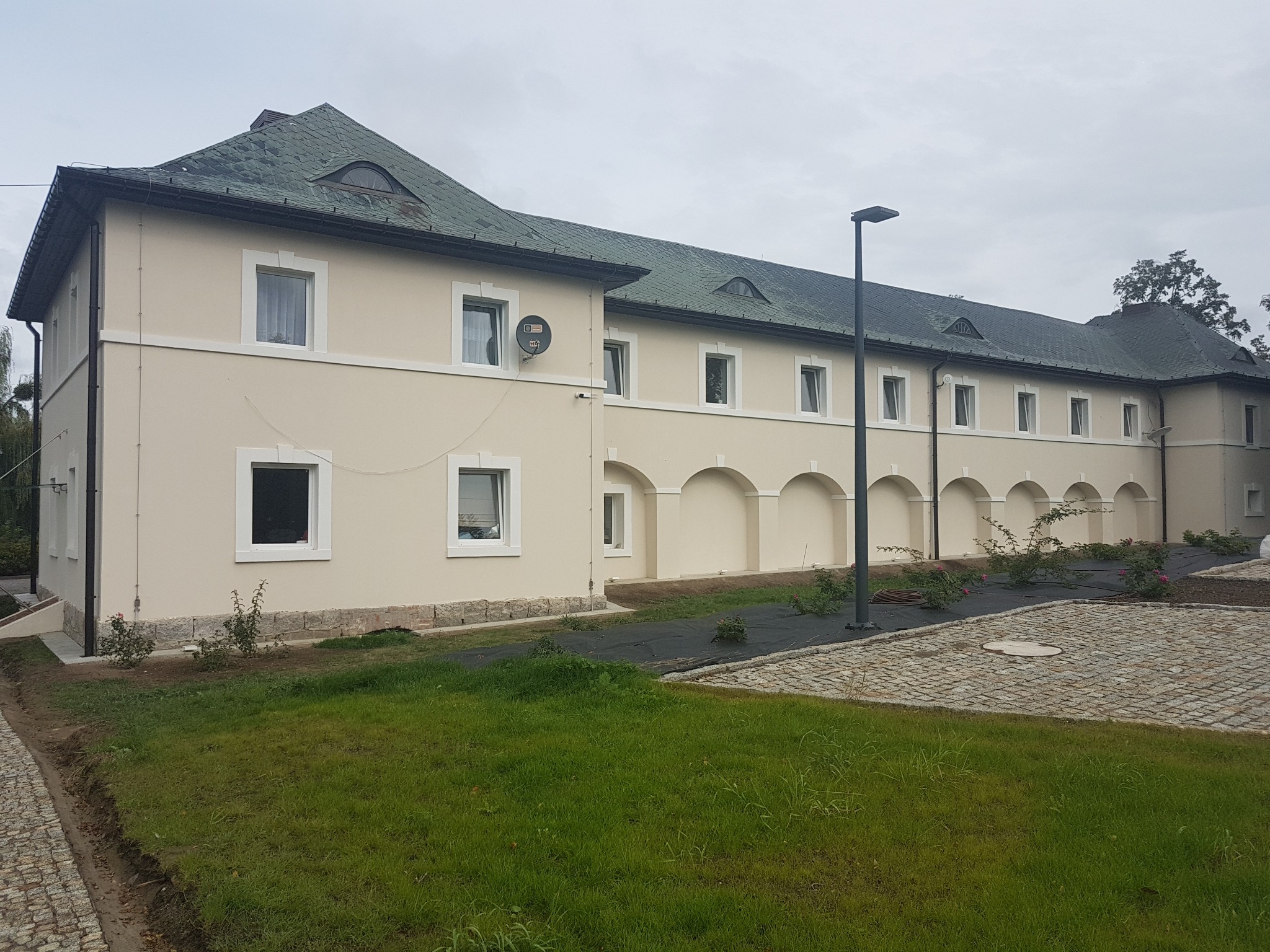 Odnowiona elewacja dwukondygnacyjnego budynku z jasną fasadą, białymi obramowaniami okien i zielonym dachem, widoczny z zewnątrz, z fragmentem trawnika i kostki brukowej.