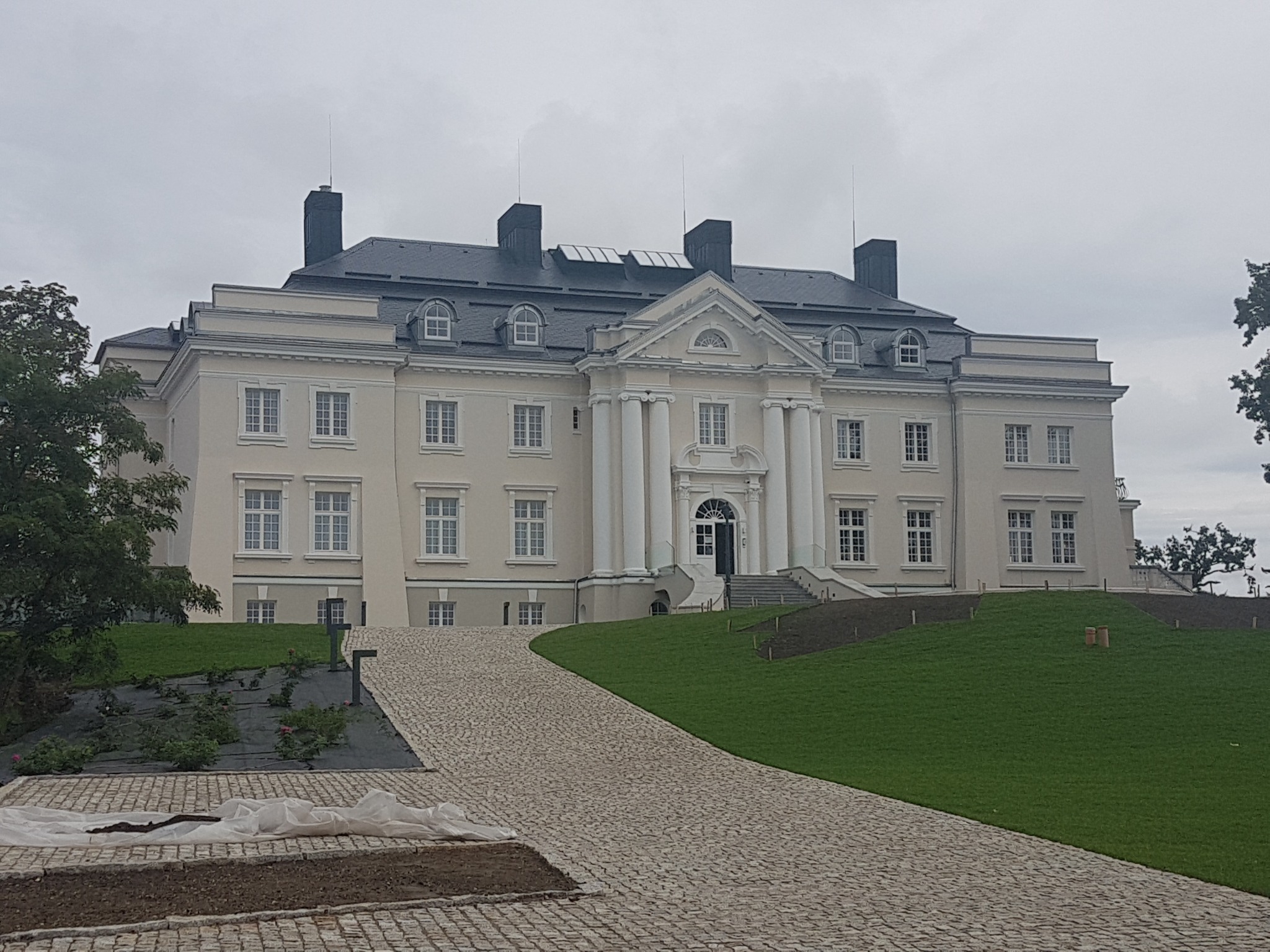 Elewacja klasycystycznego dworu po renowacji, z nową kostką brukową na podjeździe i świeżo założonym trawnikiem, widoczne elementy wykończeniowe i nasadzenia roślin.