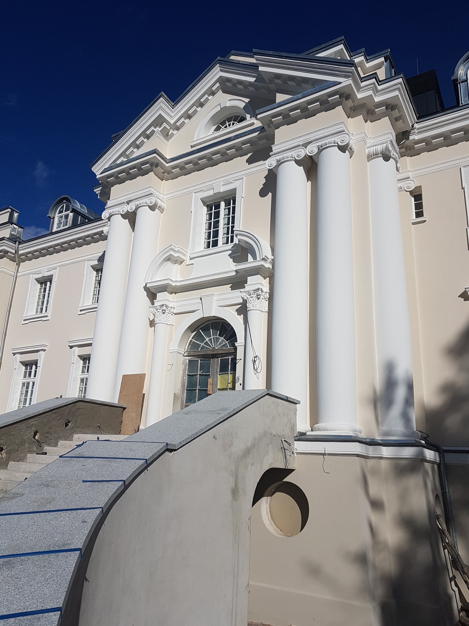 Elewacja kamienicy w trakcie renowacji, widoczne kolumny, odnowione schody wejściowe z szarego granitu i fragment rusztowania.