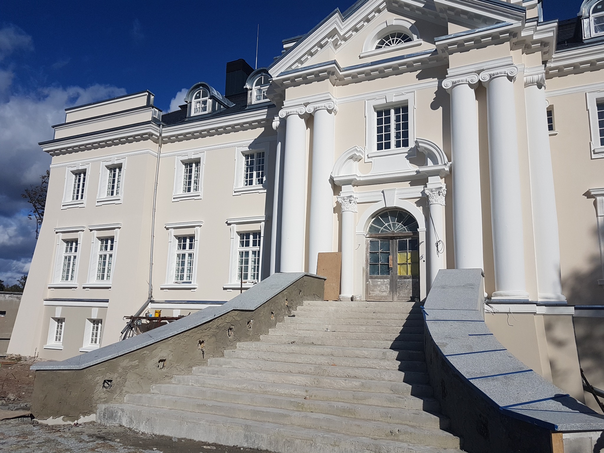 Elewacja dwukondygnacyjnego budynku z kolumnami i schodami wejściowymi w trakcie remontu, widoczne ślady prac murarskich na balustradzie schodów.