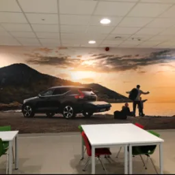Duże zdjęcie samochodu Volvo XC40 i rodziny na tle górskiego krajobrazu nadmorskiego, umieszczone na ścianie biura lub sali konferencyjnej, widoczne biurka i krzesła.