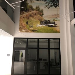 pARt - Duże zdjęcie z motywem rowerzysty i samochodu terenowego na ścianie biura, widoczne nad przeszklonymi drzwiami wejściowymi, oświetlone nowoczesnymi lampami sufitowymi.