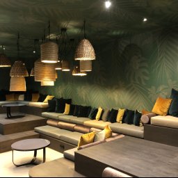 pARt - Wnętrze biura z zielonymi ścianami w liście, wielopoziomowe siedziska z poduszkami w odcieniach zieleni i żółci, wiszące lampy z naturalnego materiału.