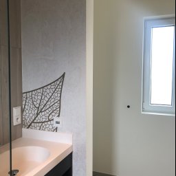 pARt - Ściana w łazience z dekoracyjną tapetą przedstawiającą motyw liścia, umywalka z szafką, białe ściany i okno w sąsiednim pomieszczeniu.