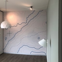 pARt - Ściana w pokoju z tapetą przedstawiającą mapę topograficzną w odcieniach bieli i niebieskiego, oświetlona przez białą lampę wiszącą o geometrycznym kształcie i lampę sufitową, z widoczną podłogą...