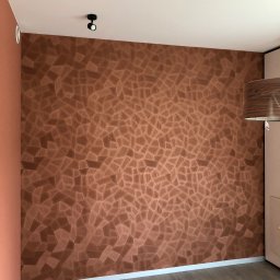 pARt - Ściana z dekoracyjną, brązową fakturą przypominającą mozaikę z drewnianych elementów, oświetlona punktowym światłem, z widocznym fragmentem szafy i podłogi.