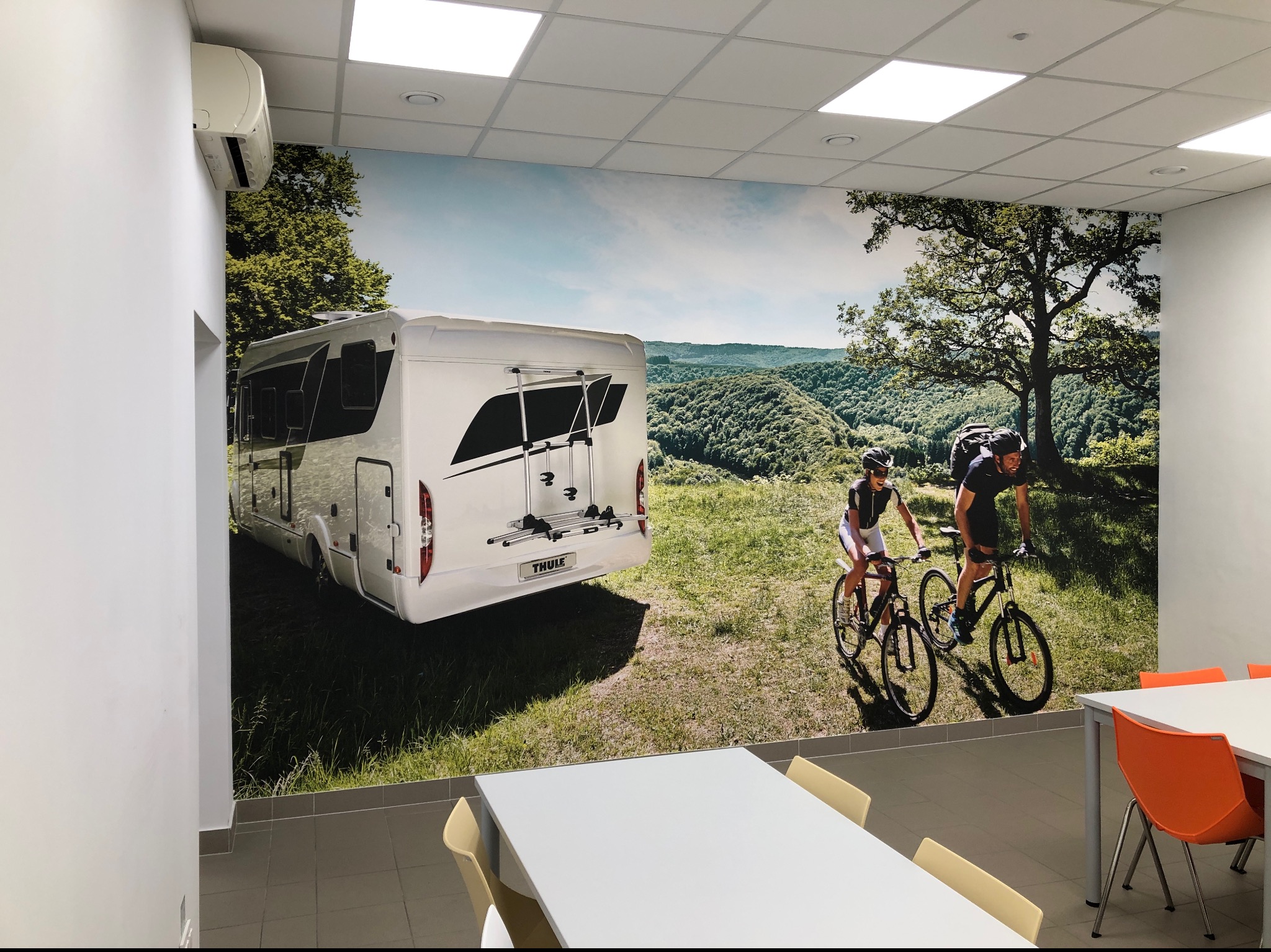 Fototapeta w biurze przedstawiająca kampera i rowerzystów na tle zielonych wzgórz, widoczna klimatyzacja na ścianie.