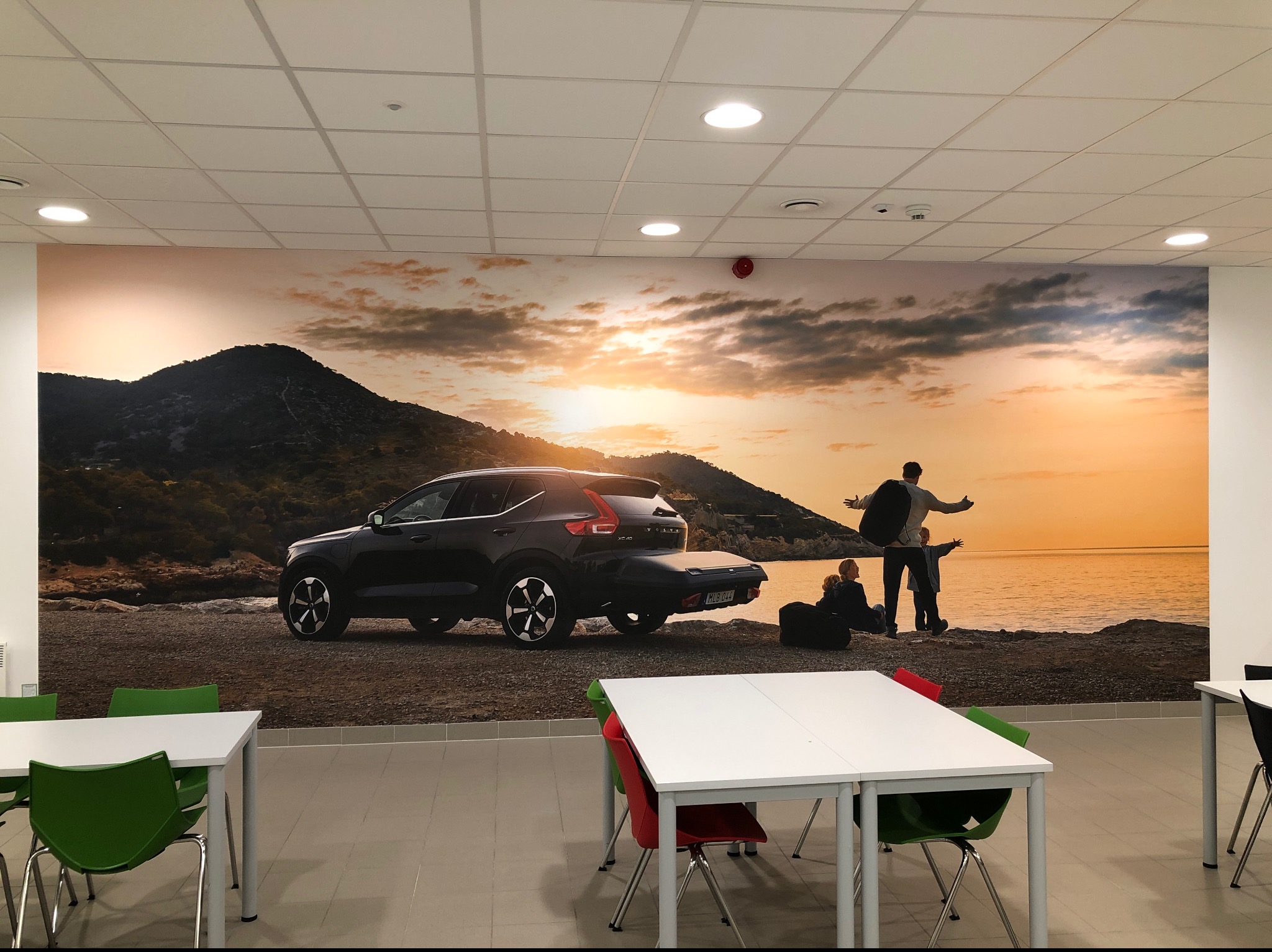 Duże zdjęcie samochodu Volvo XC40 i rodziny na tle górskiego krajobrazu nadmorskiego, umieszczone na ścianie biura lub sali konferencyjnej, widoczne biurka i krzesła.