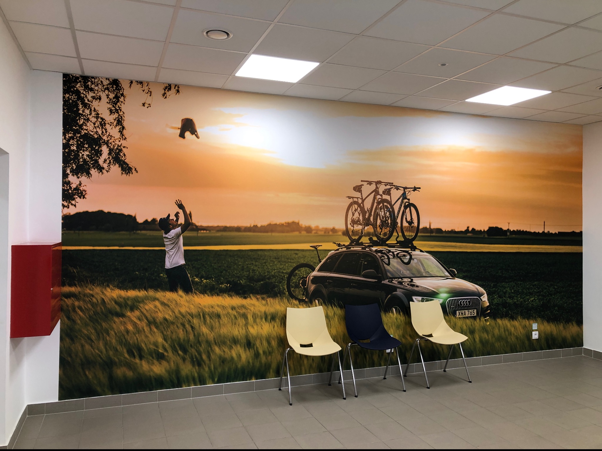Fototapeta w biurze przedstawiająca mężczyznę rzucającego czapkę w górę na tle krajobrazu z samochodem i rowerami na dachu, widoczne trzy krzesła w poczekalni.