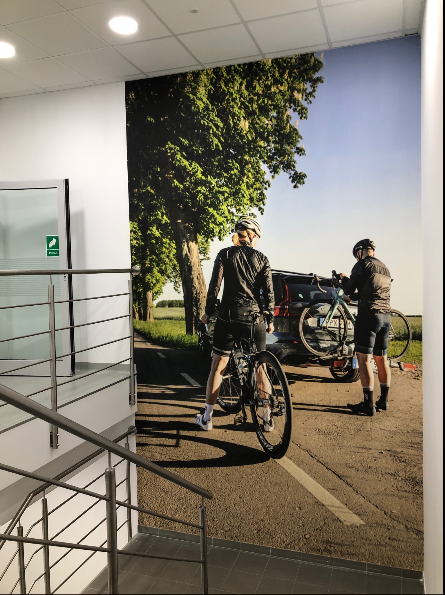 Zdjęcie przedstawia fragment ściany w biurze z fototapetą przedstawiającą parę rowerzystów przy samochodzie na tle alei drzew. W lewym dolnym rogu widoczny fragment metalowej barierki schodów.