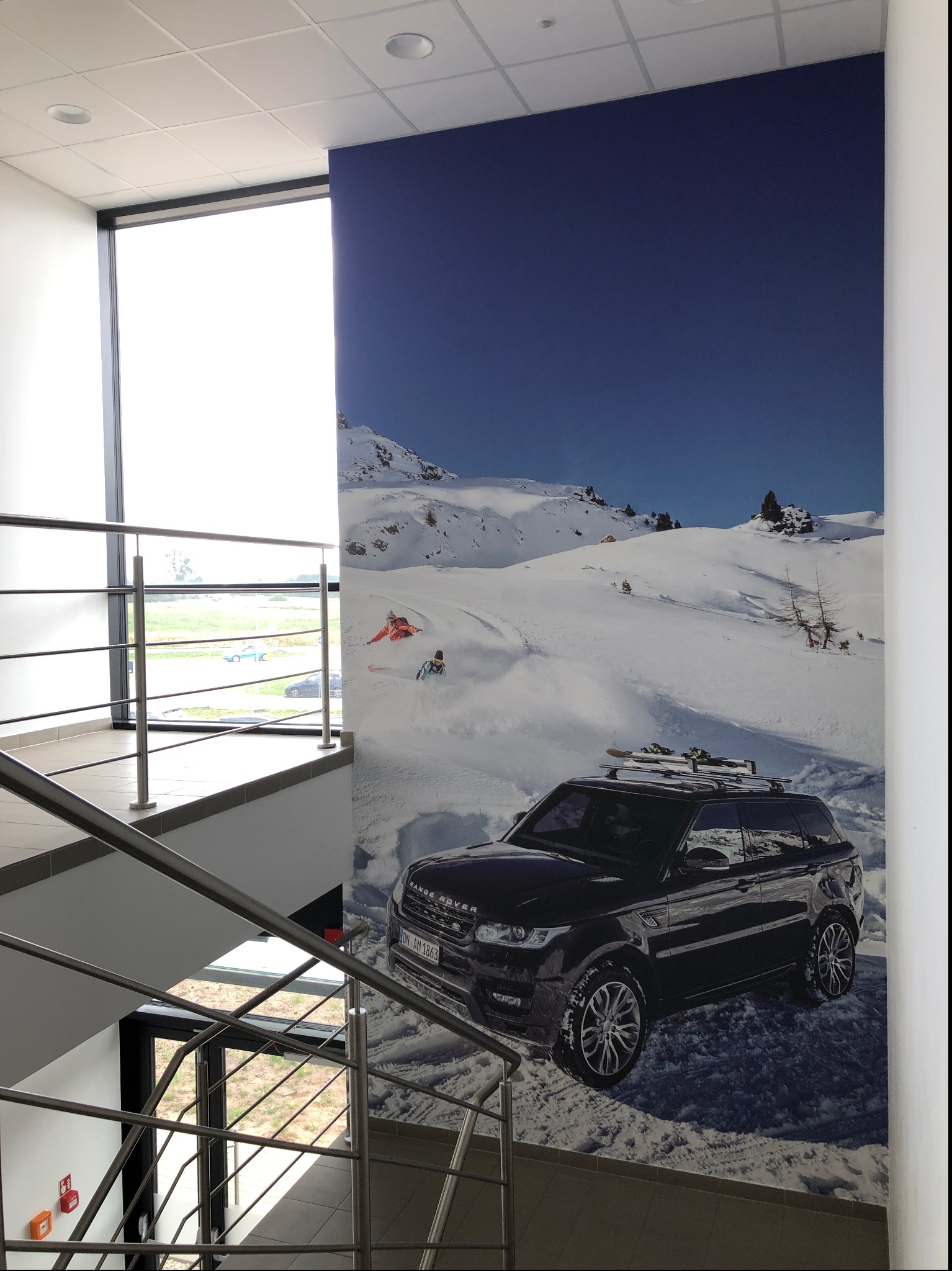 Duże zdjęcie czarnego Range Rovera z nartami na dachu, umieszczone na ścianie biura przy schodach z metalowymi barierkami, przedstawiające zimowy krajobraz z narciarzami.