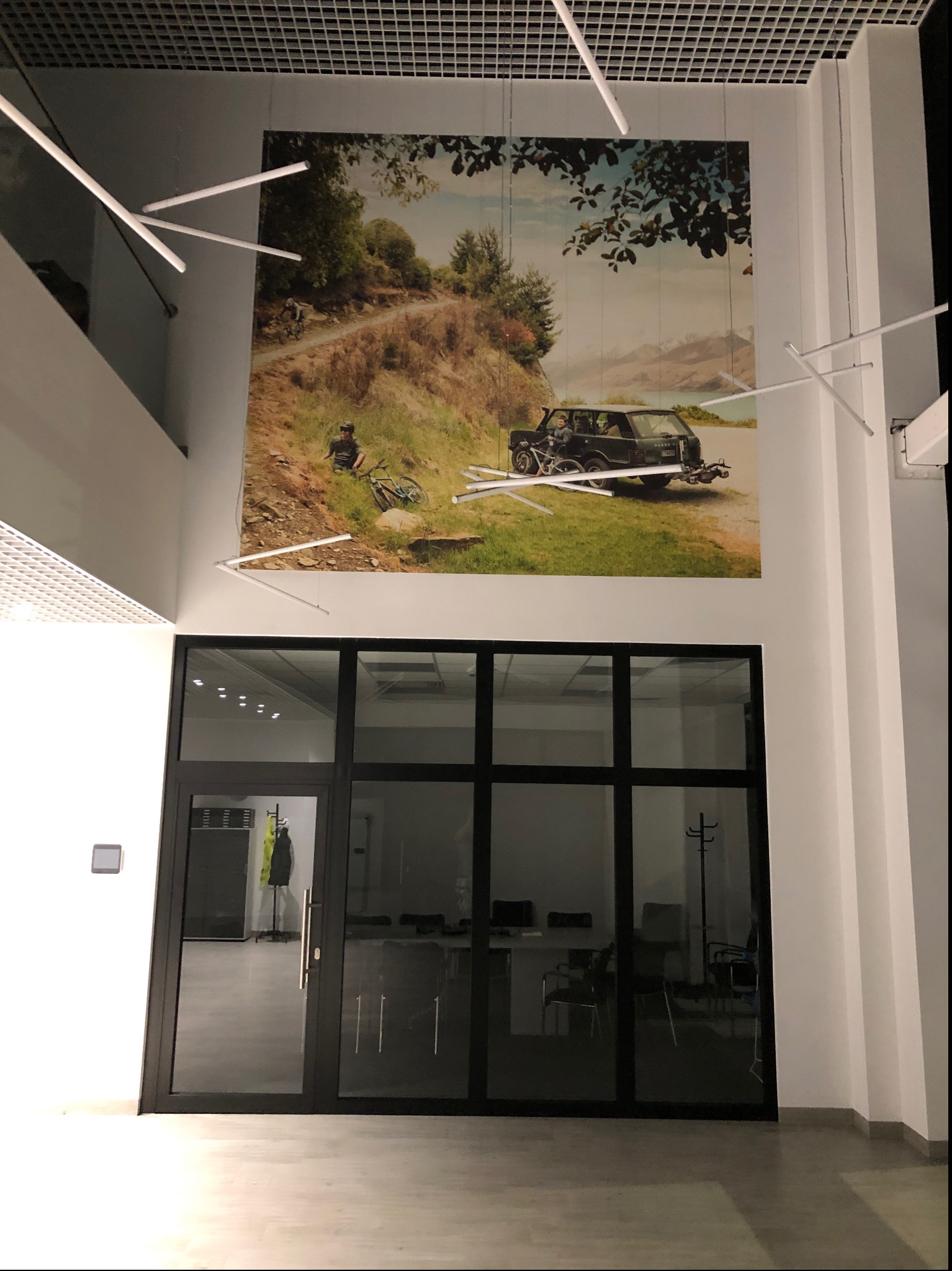 Duże zdjęcie z motywem rowerzysty i samochodu terenowego na ścianie biura, widoczne nad przeszklonymi drzwiami wejściowymi, oświetlone nowoczesnymi lampami sufitowymi.