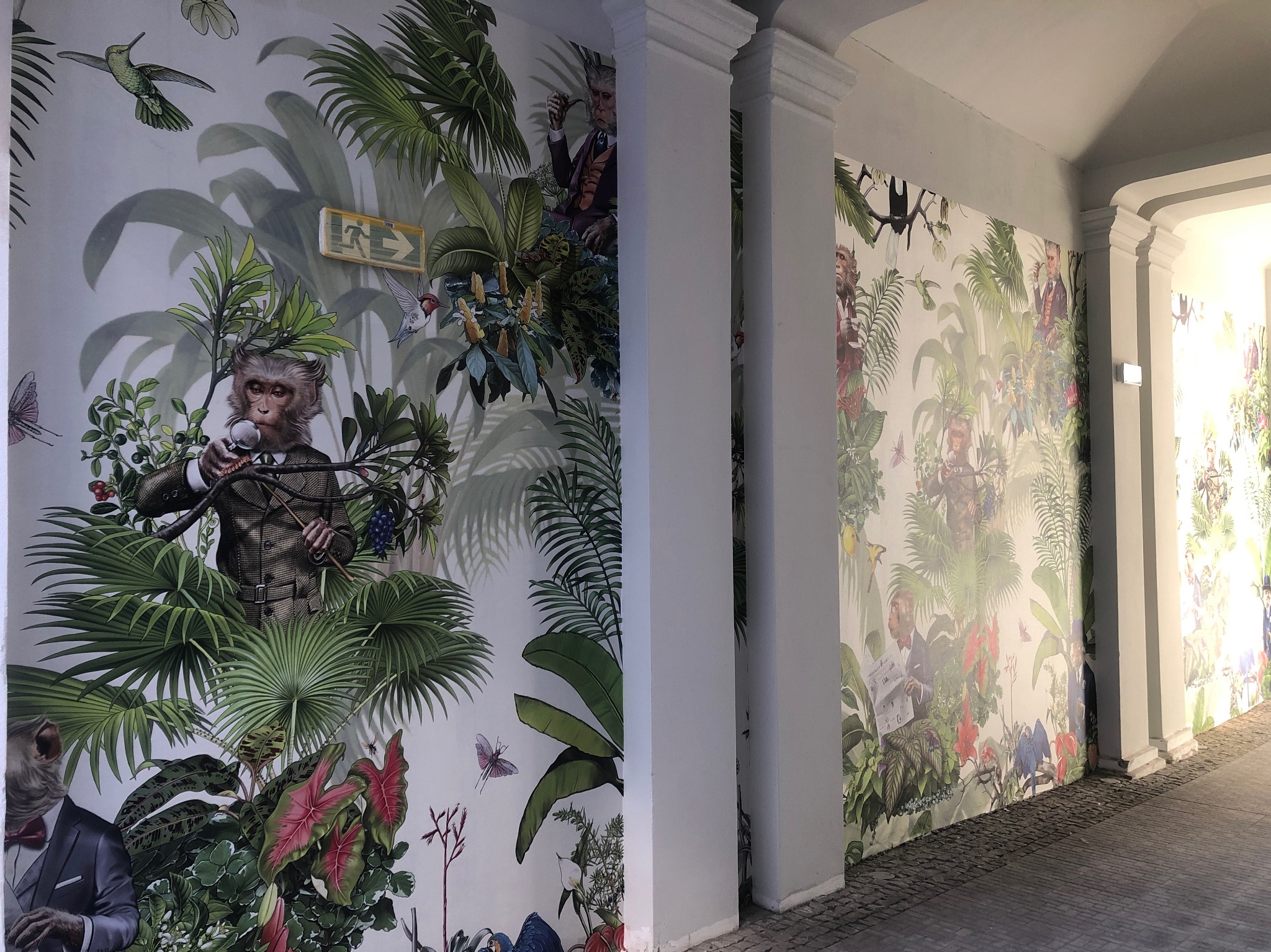 Korytarz z oryginalną tapetą przedstawiającą motywy dżungli i zwierzęta ubrane w garnitury, w tym małpę z mikrofonem i małpę czytającą gazetę, z widocznym znakiem ewakuacyjnym.