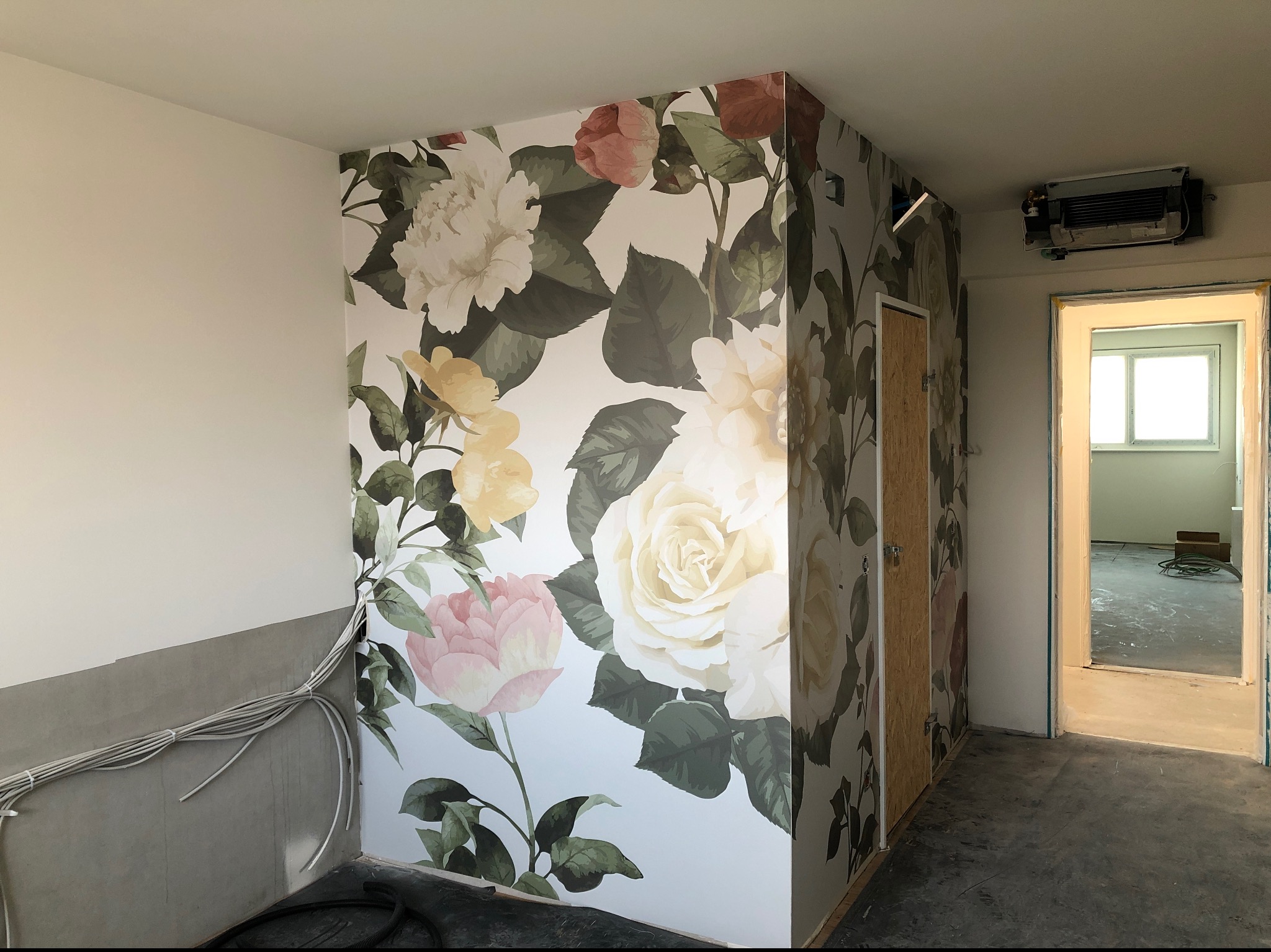 Świeżo położona tapeta z motywem kwiatowym na ścianie w pomieszczeniu w trakcie remontu, widoczne wystające kable i surowy beton na dole, oraz klimatyzator na suficie i drzwi zaklejone płytą OSB.