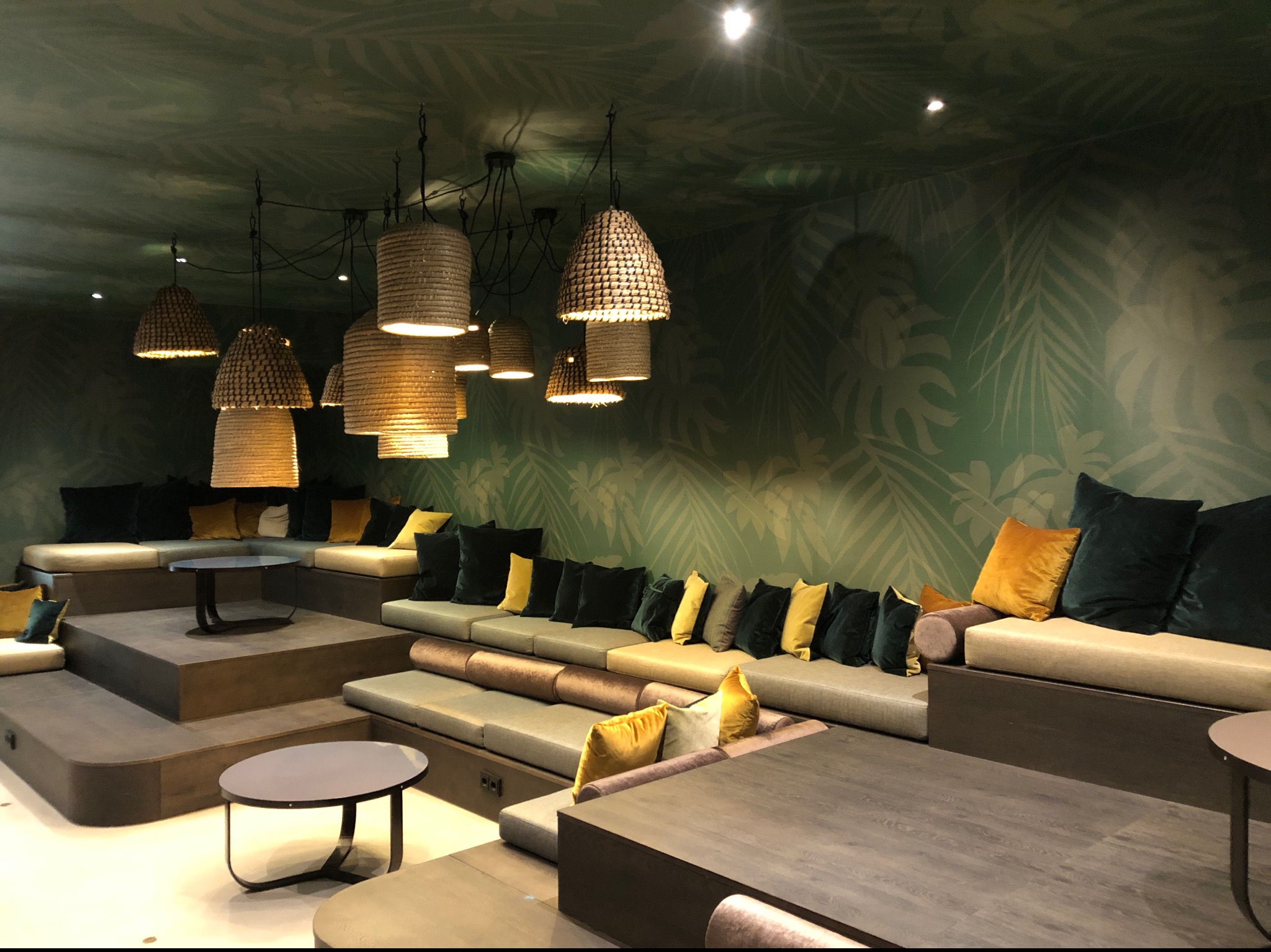 Wnętrze biura z zielonymi ścianami w liście, wielopoziomowe siedziska z poduszkami w odcieniach zieleni i żółci, wiszące lampy z naturalnego materiału.