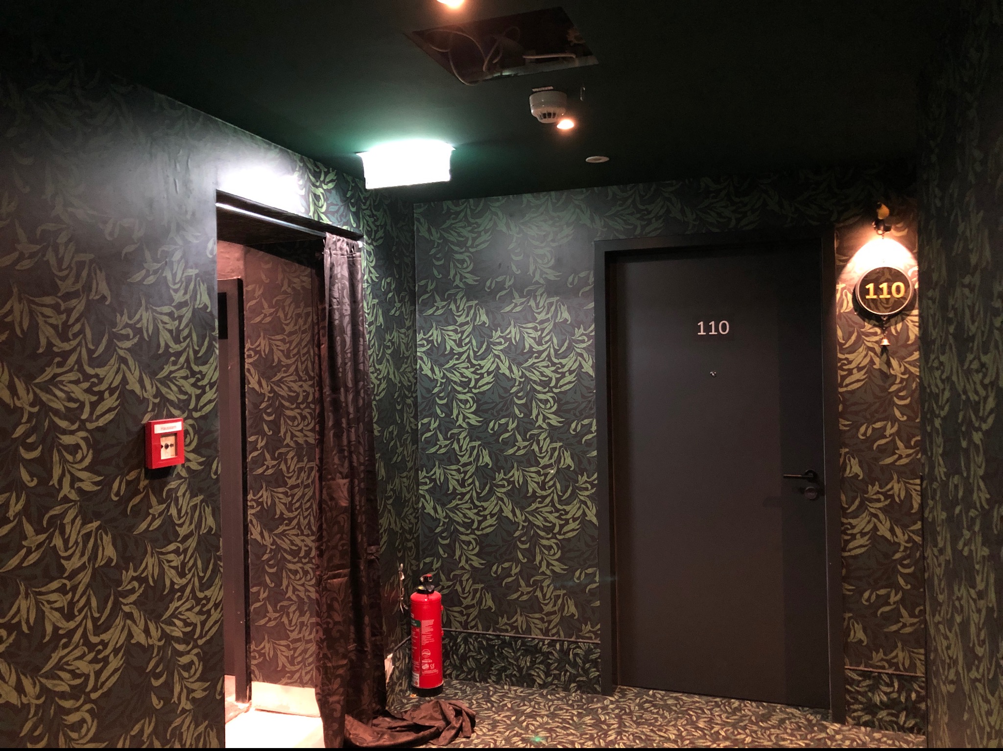 Korytarz hotelowy z ciemną, wzorzystą tapetą w liście, numerem pokoju 110 nad drzwiami, gaśnicą i zasłoną przy wejściu.