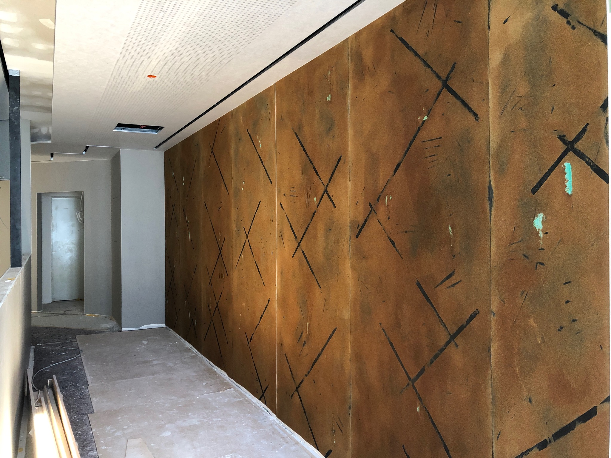 Brązowa ściana z geometrycznym wzorem w trakcie malowania w biurze, widoczne ślady farby i niedokończone wykończenie, sufit podwieszany z oświetleniem.