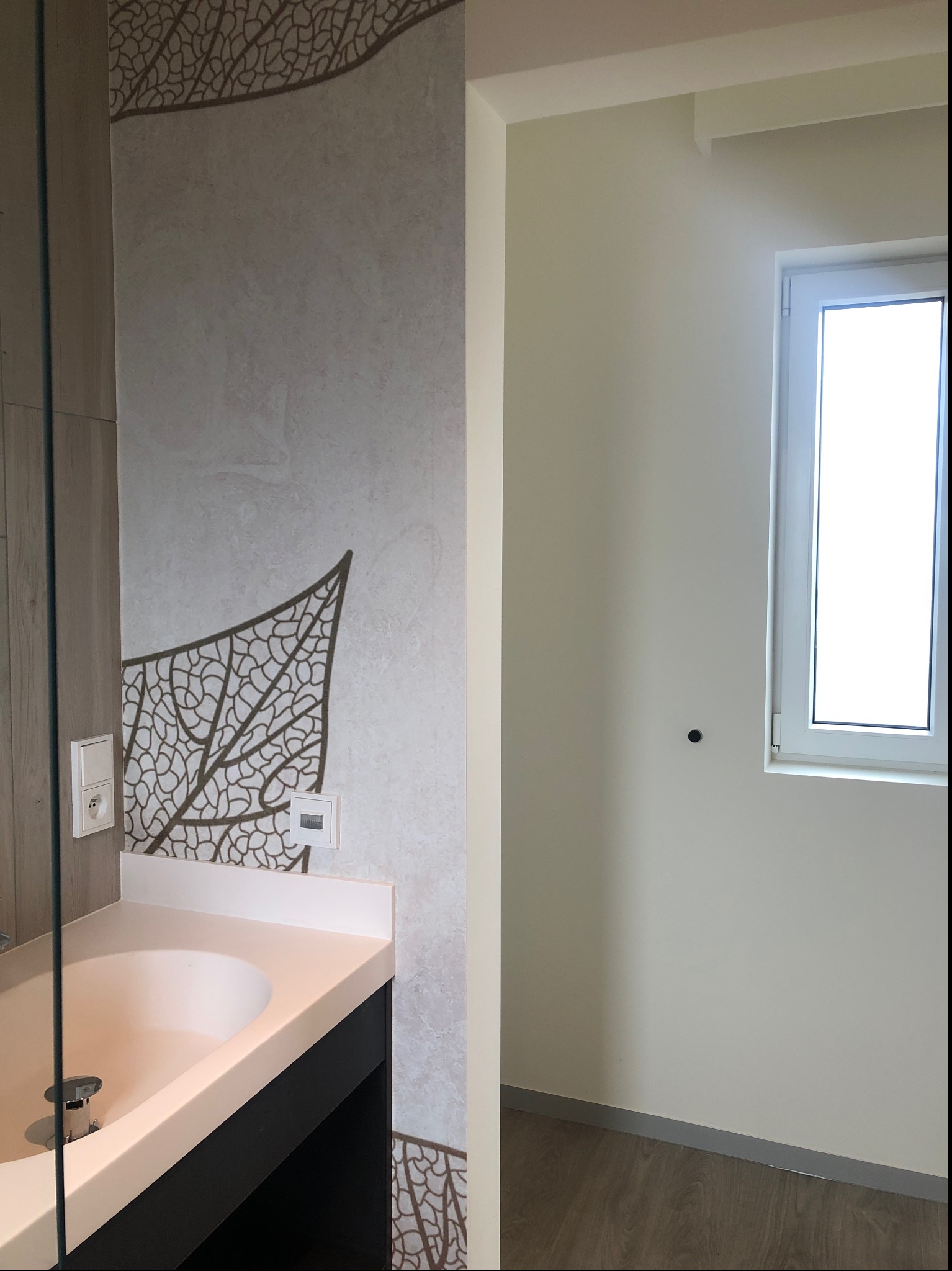 Ściana w łazience z dekoracyjną tapetą przedstawiającą motyw liścia, umywalka z szafką, białe ściany i okno w sąsiednim pomieszczeniu.